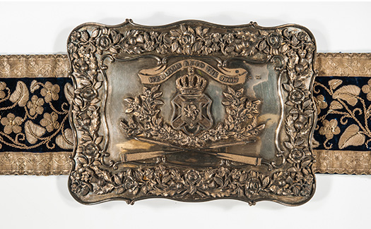 Image of a decorative belt and buckle made of silver, velvet, leather. The buckle with the motto wreathed in mayflowers: “We bloom amid the snow.”