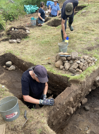 An outdoor, high-angle shot of an active archaeological excavation site on a grassy field. In the foreground, Dr. Katie Cottreau-Robins, wearing a dark purple baseball cap, a black t-shirt, and patterned work gloves stands inside a deeply excavated, rectangular dirt trench, carefully clearing soil from a dirt wall. Next to the trench sits a metal bucket and a small trowel on the grass.
