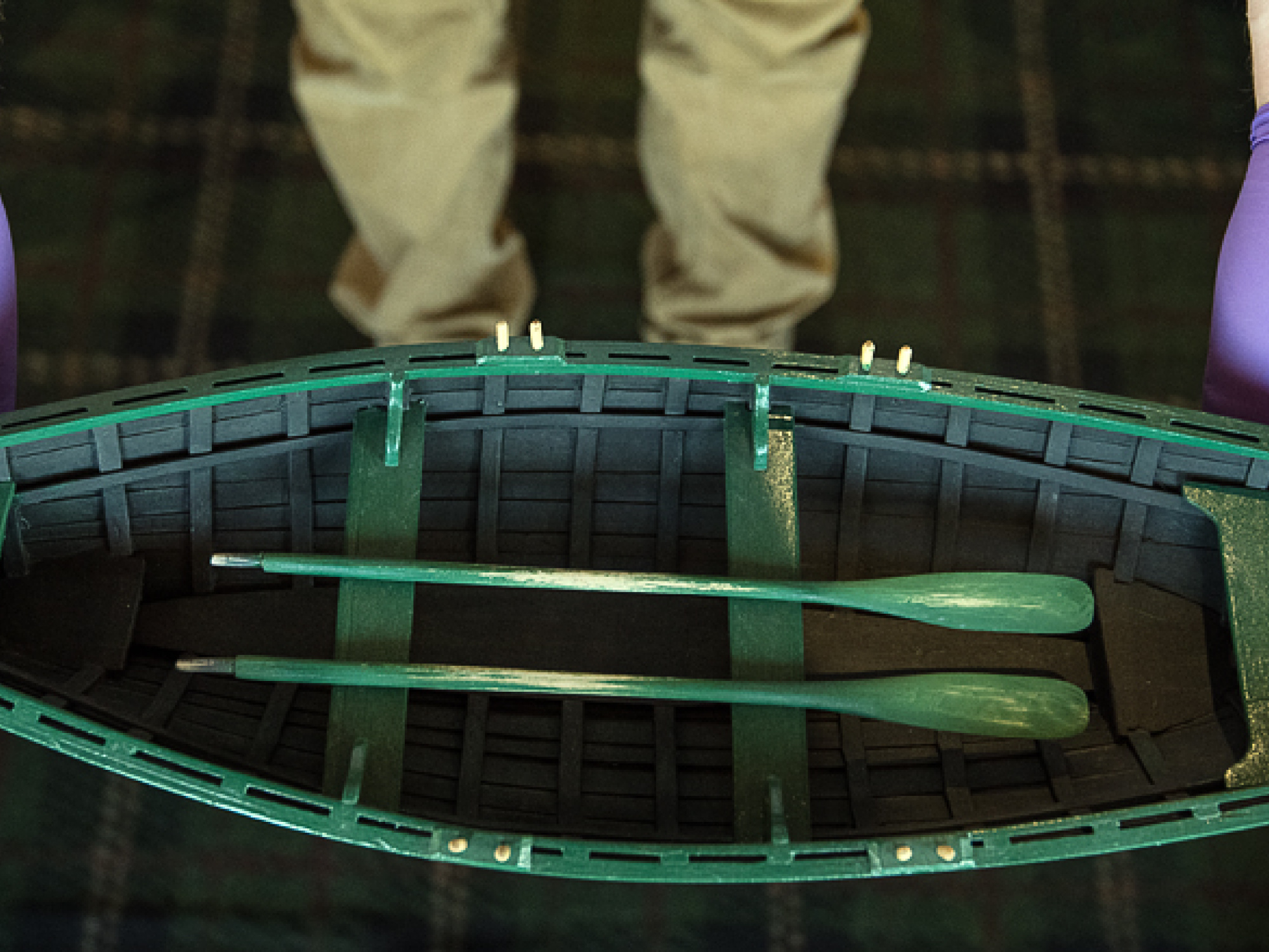 A close-up, overhead shot of a person wearing purple nitrile gloves carefully holding a detailed miniature model of a green wooden dory. The model boat features tiny green oars resting inside the hull. The background shows the person's khaki trousers and a dark, patterned carpet.
