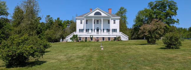 Uniacke Estate a grand white house with a classical design, including a columned entrance and a long veranda. The house is surrounded by a well-maintained lawn and mature trees.