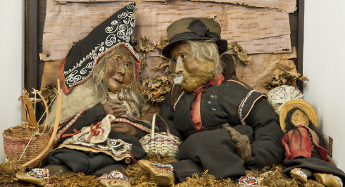  A diorama featuring two life-sized, elderly Mi'kmaq figures seated against a backdrop of birch bark, possibly representing a couple. The woman on the left wears a pointed black cap (likely a tuwaqan) adorned with white beaded floral patterns, and a dark cloth garment with contrasting trim. The man on the right wears a dark felt hat and a dark coat, holding a pipe. Both figures have realistic, aged faces, hands, and long grey hair.