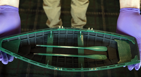 A close-up, overhead shot of a person wearing purple nitrile gloves carefully holding a detailed miniature model of a green wooden dory. The model boat features tiny green oars resting inside the hull. The background shows the person's khaki trousers and a dark, patterned carpet.