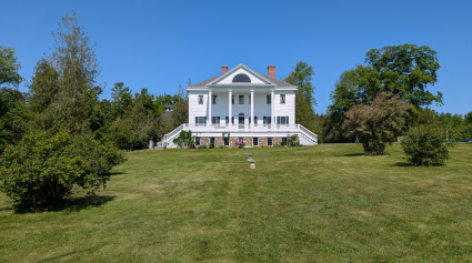 Uniacke Estate a grand white house with a classical design, including a columned entrance and a long veranda. The house is surrounded by a well-maintained lawn and mature trees.