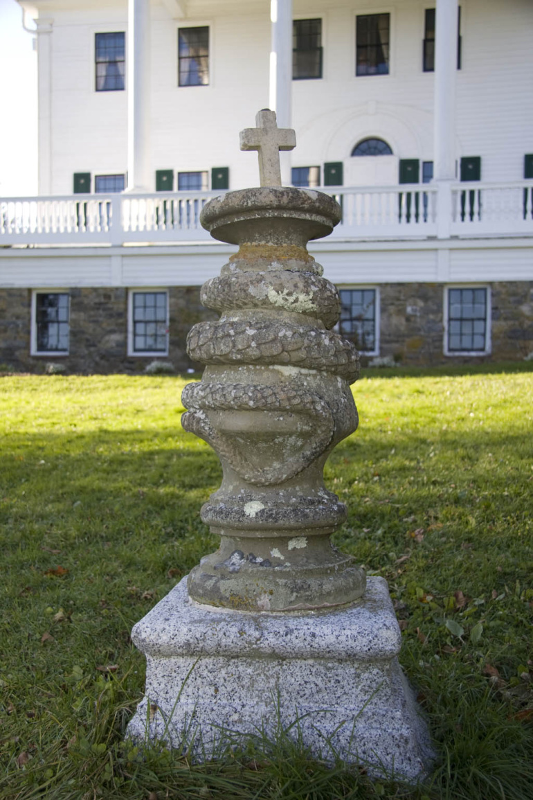 A stone monument with a cross on top, standing in front of a large white house.