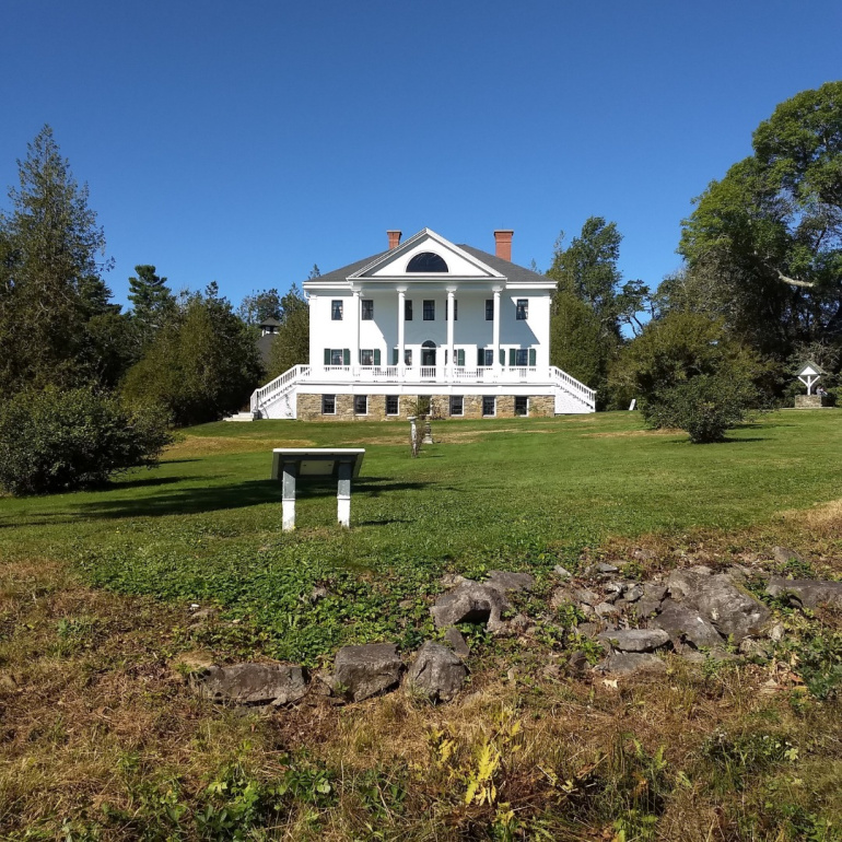 Uniacke Estate a grand white house with a classical design, including a columned entrance and a long veranda. The house is surrounded by a well-maintained lawn and mature trees.