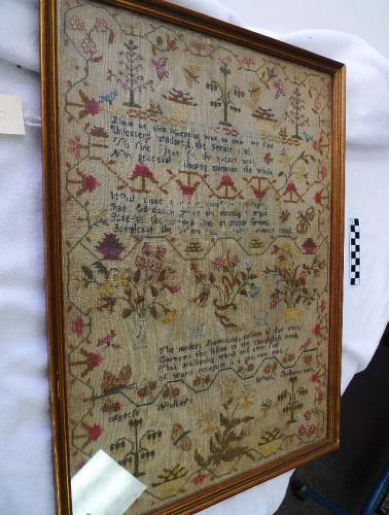 A framed antique needlework sampler under glass, displayed at an angle against a white cloth backdrop. The rectangular sampler features rows of stitched text interspersed with various decorative motifs, including stylized trees, birds, butterflies, baskets, and floral arrangements in muted red, green, blue, and yellow threads on a darkened linen fabric. A narrow gold-colored wooden frame encloses the textile.
