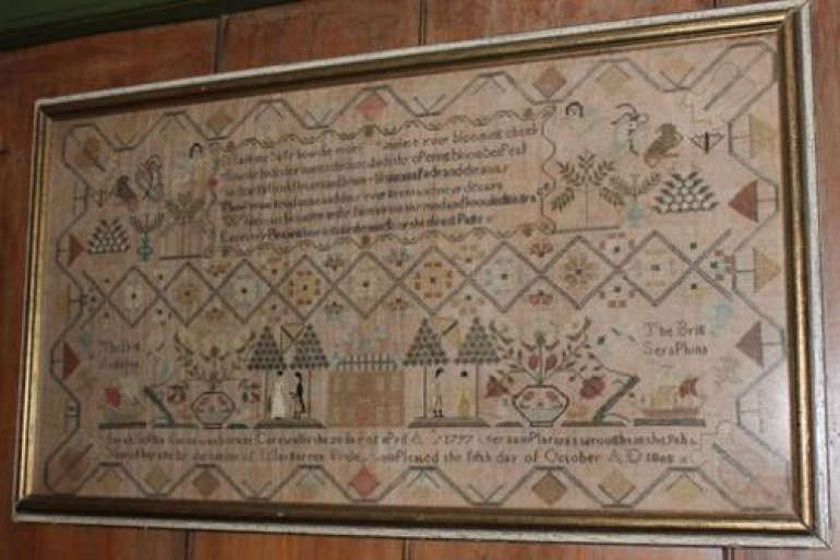 A wide, rectangular antique needlework sampler mounted in a gold-edged wooden frame against a wood-paneled wall. The intricately stitched sampler features a complex geometric diamond-patterned border enclosing rows of text, floral vases, stylized trees, and animal figures. Along the lower section, a detailed landscape scene depicts a building flanked by small human figures, trees, and sailing ships, with an inscription stitched across the bottom edge.