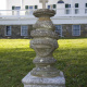 A stone monument with a cross on top, standing in front of a large white house.