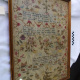 A framed antique needlework sampler under glass, displayed at an angle against a white cloth backdrop. The rectangular sampler features rows of stitched text interspersed with various decorative motifs, including stylized trees, birds, butterflies, baskets, and floral arrangements in muted red, green, blue, and yellow threads on a darkened linen fabric. A narrow gold-colored wooden frame encloses the textile.