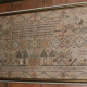 A wide, rectangular antique needlework sampler mounted in a gold-edged wooden frame against a wood-paneled wall. The intricately stitched sampler features a complex geometric diamond-patterned border enclosing rows of text, floral vases, stylized trees, and animal figures. Along the lower section, a detailed landscape scene depicts a building flanked by small human figures, trees, and sailing ships, with an inscription stitched across the bottom edge.