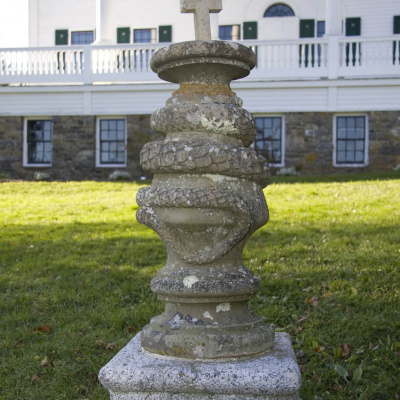 A stone monument with a cross on top, standing in front of a large white house.