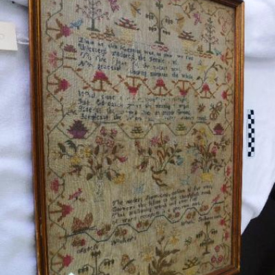 A framed antique needlework sampler under glass, displayed at an angle against a white cloth backdrop. The rectangular sampler features rows of stitched text interspersed with various decorative motifs, including stylized trees, birds, butterflies, baskets, and floral arrangements in muted red, green, blue, and yellow threads on a darkened linen fabric. A narrow gold-colored wooden frame encloses the textile.