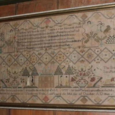 A wide, rectangular antique needlework sampler mounted in a gold-edged wooden frame against a wood-paneled wall. The intricately stitched sampler features a complex geometric diamond-patterned border enclosing rows of text, floral vases, stylized trees, and animal figures. Along the lower section, a detailed landscape scene depicts a building flanked by small human figures, trees, and sailing ships, with an inscription stitched across the bottom edge.