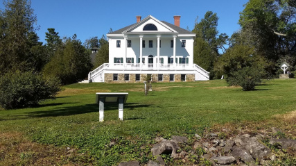 Uniacke Estate a grand white house with a classical design, including a columned entrance and a long veranda. The house is surrounded by a well-maintained lawn and mature trees.
