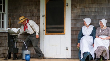 Two female costumed interpreters sit on the porch of Rosebank cottage watching a male costumed interpreter churn butter.