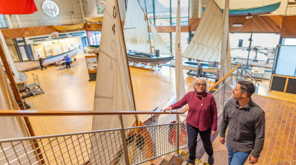 A woman and a man are descending a wide, tiled staircase with metal railings inside a large, bright museum hall. The woman, with short white hair and glasses, is wearing a red sweater and black pants. The man, with short dark hair, is wearing a dark grey shirt and jeans. Surrounding them are several historical wooden sailboats with white sails, some hanging from the ceiling and others displayed on the floor below. The background shows more exhibits, including small boats and display cases.