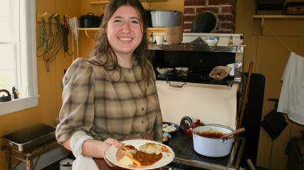 A historical interpreter in a vintage kitchen holds a plate of traditional baked beans.