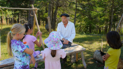 An older man in historical attire, wearing a loose white shirt and a black flat cap, sits on a rustic wooden bench in a grassy, wooded clearing. He is speaking to four young children gathered around him: two girls on the left, one in the center wearing a colorful patterned sun hat with her back to the camera, and one on the right in a yellow t-shirt.