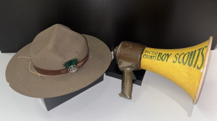 On the left, a classic olive-drab felt Boy Scout campaign hat. On the right, an old-fashioned portable megaphone sits on another small black block. It has a metallic, olive-green handle and housing, attached to a bright yellow cone-shaped horn. Hand-painted in bold, dark green letters on the side of the yellow cone is the text: "PICTOU COUNTY BOY SCOUTS"