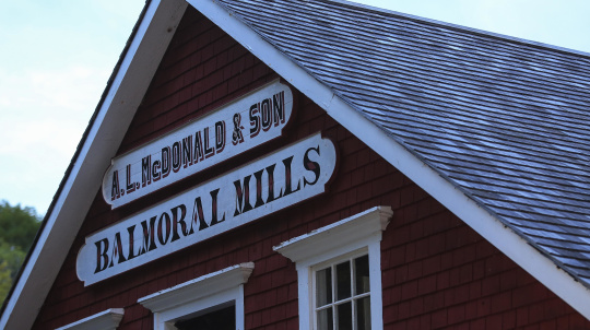 A close-up shot of the Balmoral Mills sign, featuring white lettering on a dark background, attached to the red shingle siding of a building. The sign reads 'A.L. McDonald & Son' above 'Balmoral Mills'. A portion of the building's gray shingle roof and a white-framed window are also visible.