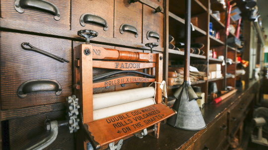 A close-up view of a vintage washing machine wringer, labeled 'FALCON,' with wooden rollers and metal components. The wringer is positioned in front of a multi-drawer wooden cabinet with small, dark metal handles. The drawers and other shelves in the background are filled with various unidentified objects.