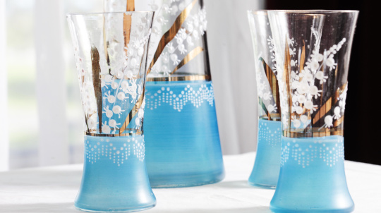 Four tall, clear glass vases with blue bases and white floral patterns, arranged on a white tablecloth. Two vases are in front, and two are in the background.