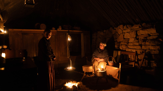 Costumed interpreters, two women by a fire at night at Baile nan Gàidheal | Highland Village. 