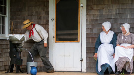 Two female costumed interpreters sit on the porch of Rosebank cottage watching a male costumed interpreter churn butter.