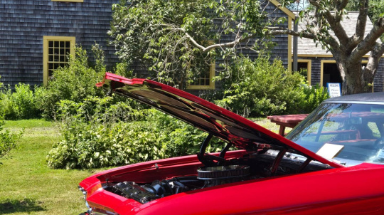 A classic red muscle car on display at the North Hills Show & Shine, with the house building behind it.