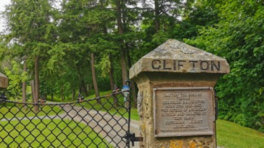  An outdoor photograph showing a stone gatepost with an ornate metal gate partially open on the left. The gatepost is made of rough-hewn stone and has the word "CLIFTON" carved into the top. Below the carved word is a bronze plaque, which is difficult to read but appears to commemorate General Charles Rainsford, born 1782. died 1855The gate opens onto a path leading uphill into a wooded area with tall trees and lush greenery.