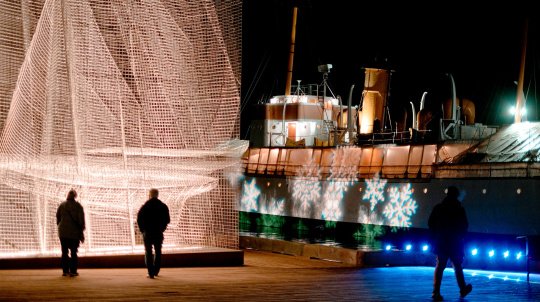  An outdoor night shot. Two people walk away from the camera along a wooden pier toward a large, illuminated, conical sculpture made of fine, reflective mesh on the left. To the right, the CSS Acadia is docked, its hull partially illuminated by projected white snowflake patterns.
