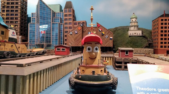  A large model of Theodore Tugboat, the main character from the Canadian children's television show, is displayed in a miniature version of a city harbor. Theodore is a smiling tugboat with a red cap. The background features models of various buildings, including a prominent hill with a white clock tower (resembling the Halifax Citadel Clock Tower), suggesting the setting is based on Halifax, Nova Scotia.