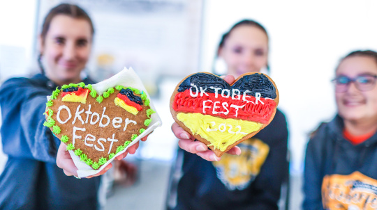  Two people hold up heart-shaped gingerbread cookies decorated for Oktoberfest, while a third person smiles in the background.