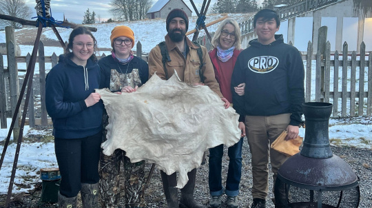 Five people stand outdoors in a snowy landscape, smiling and holding up a large, light-colored animal hide. They are dressed in winter clothing, including beanies, hoodies, and boots; one person wears camouflage overalls. In the background, there is a wooden fence, a white barn, and a few bare trees under an overcast sky. A black metal fire pit sits in the lower right corner of the frame.