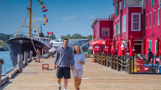 A smiling couple walks hand-in-hand along a wooden boardwalk, flanked by a large ship named "Cape Sable" on one side and vibrant red buildings on the other