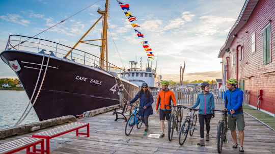 A group of four cyclists walk their bikes along a wooden boardwalk at sunset, passing the large black-hulled ship "Cape Sable" and a bright red building.