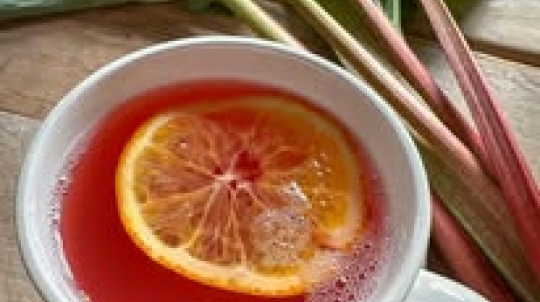 A white ceramic mug filled with a bright pink rhubarb beverage, garnished with a floating slice of orange. The mug sits on a wooden surface next to several fresh stalks of rhubarb with long red and green stems and large green leaves.