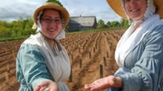 Two smiling women dressed in historical 19th-century attire stand in the foreground of a large, freshly tilled agricultural field under a blue sky. Both women wear long blue dresses with white aprons, white bonnets, and wide-brimmed straw hats. They hold out their open hands toward the camera, displaying small seeds palm-up. In the background, rows of dirt mounds stretch across the field toward a grey wooden house and distant green trees.