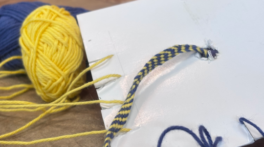 An image showing a close-up, high-angle view of a handmade cardboard loom with a partially woven blue and yellow friendship bracelet.