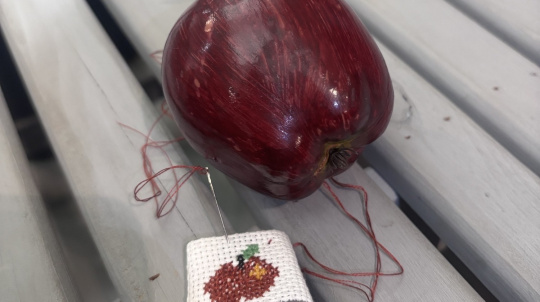 An image showing a high-angle view of a deep red artificial apple and a small cross-stitch project resting on a light grey, slatted wooden surface.