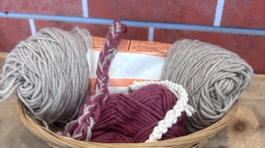 An eye-level, close-up shot of a small, woven bamboo basket filled with skeins of yarn and handmade braided cords, sitting on a rustic wooden table against a red brick background.