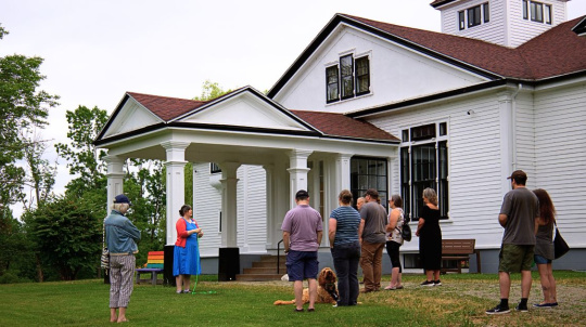A tour guide in a bright blue dress and orange cardigan addresses a small group of visitors during an outdoor history walk on the grassy lawn of a historic white estate. The large building features classic white siding, dark window trim, and a prominent columned portico over the entrance. A golden-brown dog rests on the grass next to one of the visitors, and a colorful rainbow-painted bench is visible in the background under a soft, overcast sky.